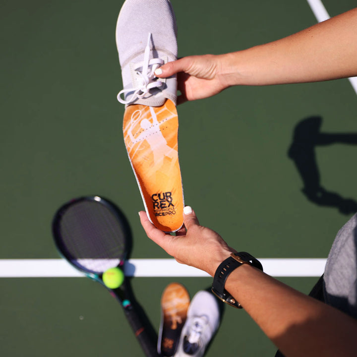 Person standing on green tennis court while inserting CURREX ACEPRO yellow medium profile insoles into gray tennis shoe #1-wahle-dein-profil_med