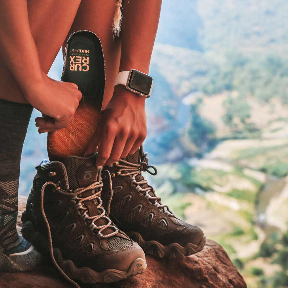 Woman placing yellow medium profile CURREX HIKEPRO insoles into hiking shoes while standing near mountain cliff #1-wahle-dein-profil_med